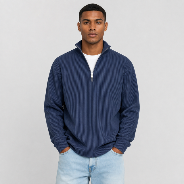 A man wearing a navy blue ribbed quarter-zip pullover sweater over a white t-shirt and light blue jeans, standing against a light gray studio background.