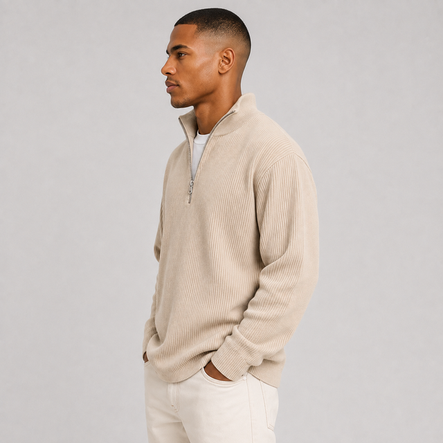 A man in profile wearing a ribbed beige half-zip sweater over a white undershirt and white pants, standing against a plain light gray studio background.