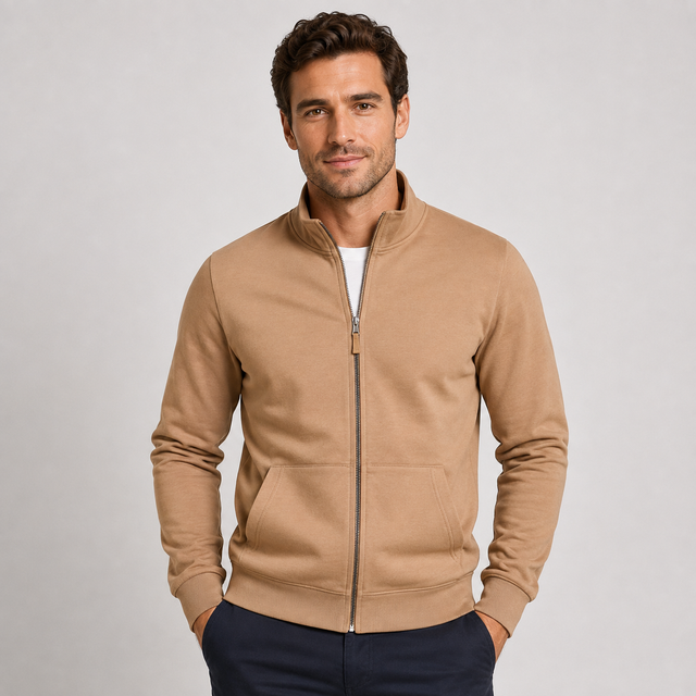 A man wearing a tan zip-up fleece jacket with a stand collar and front pockets, paired with dark navy pants, standing against a light gray studio background.