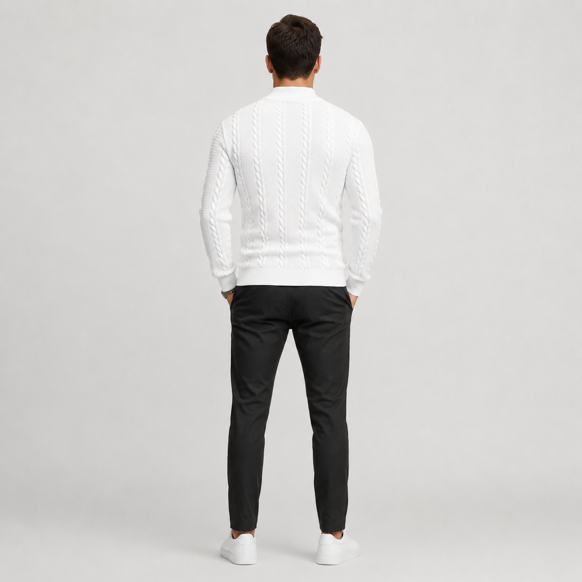 A man viewed from behind wearing a white cable-knit mock-neck sweater, black trousers, and white sneakers against a light gray studio background.
