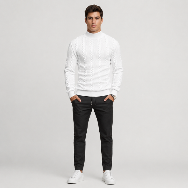 A man modeling a white cable-knit mock neck sweater, paired with black drawstring trousers and white sneakers, standing against a light gray studio background.