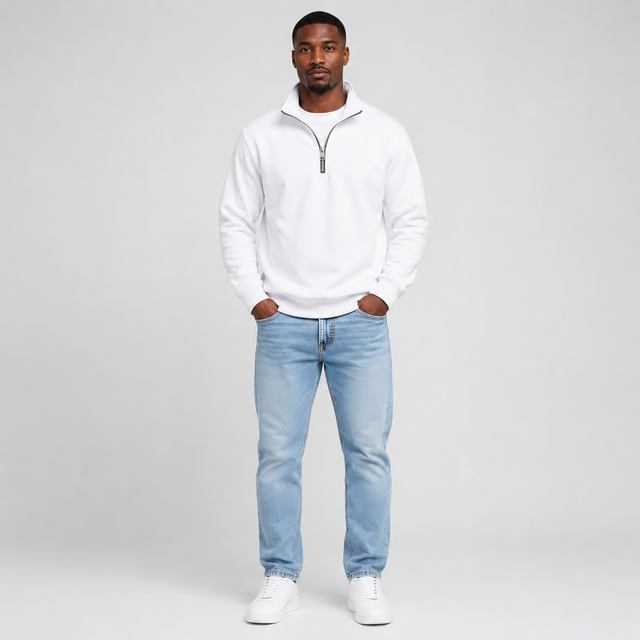 A man modeling a white half-zip pullover sweatshirt with light blue jeans and white sneakers, standing against a light gray studio background.