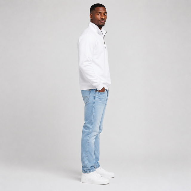 A man standing in profile wearing a white quarter-zip pullover, light blue jeans, and white sneakers against a light gray studio background.