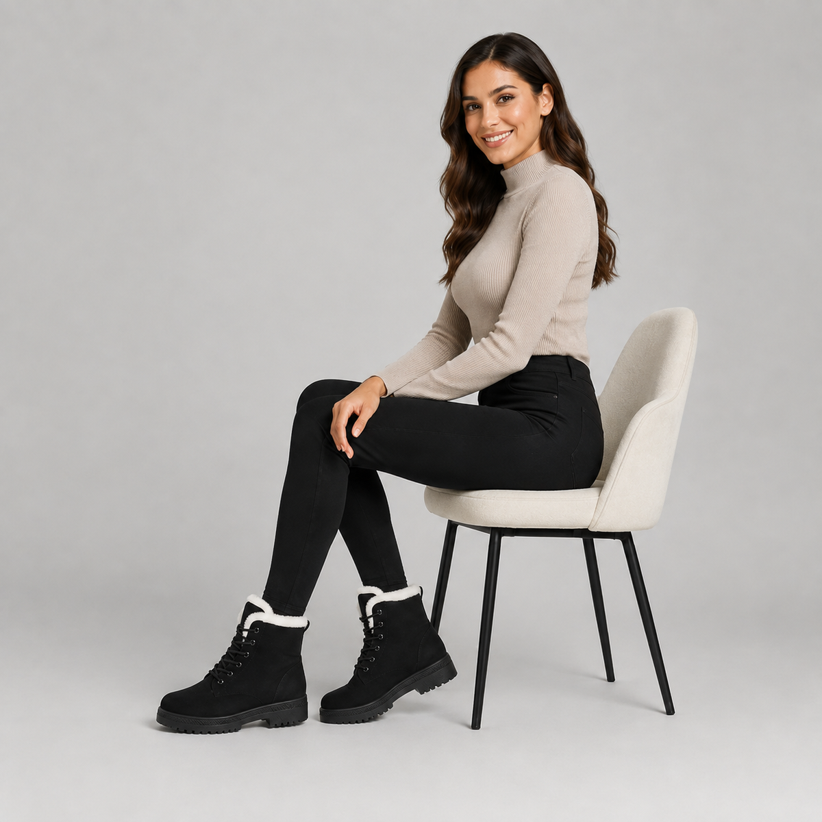 A woman sits on a light beige chair wearing a beige mock-neck long-sleeve top, black pants, and black lace-up boots with white fleece lining, against a solid light gray background.