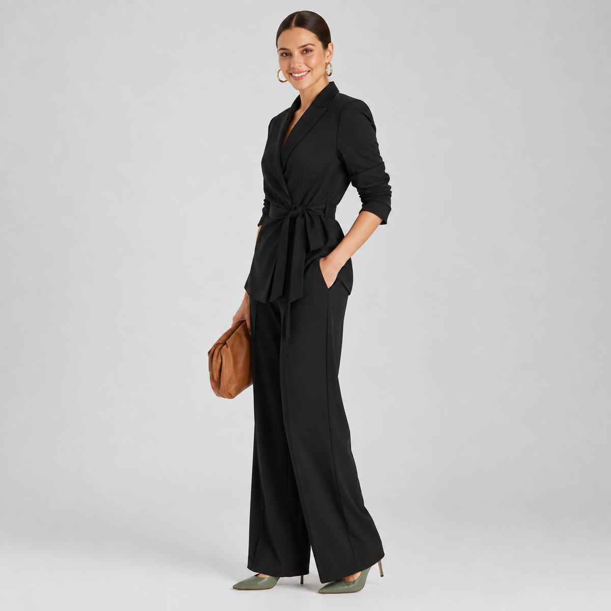A woman modeling a black belted wrap-style jumpsuit with wide legs, paired with sage green heels and a brown clutch, against a light gray studio background.