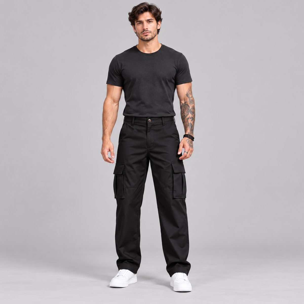 A man modeling black cargo pants with a black t-shirt and white sneakers, standing against a solid gray studio background.