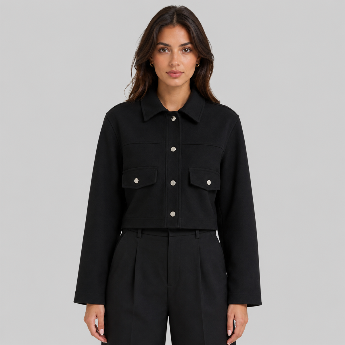 A woman modeling a black cropped button-front jacket with flap chest pockets and matching black trousers, photographed against a plain light gray studio background.