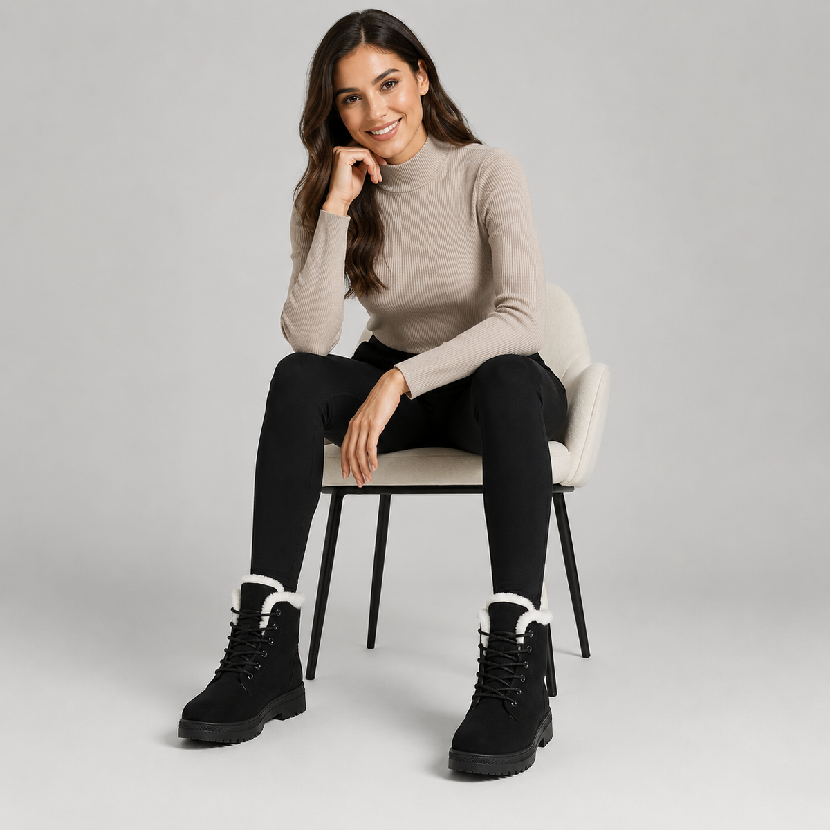 A woman sits on a light gray chair wearing a beige mock-neck knit sweater, black pants, and black lace-up boots with white faux-fur trim, against a light gray studio background.