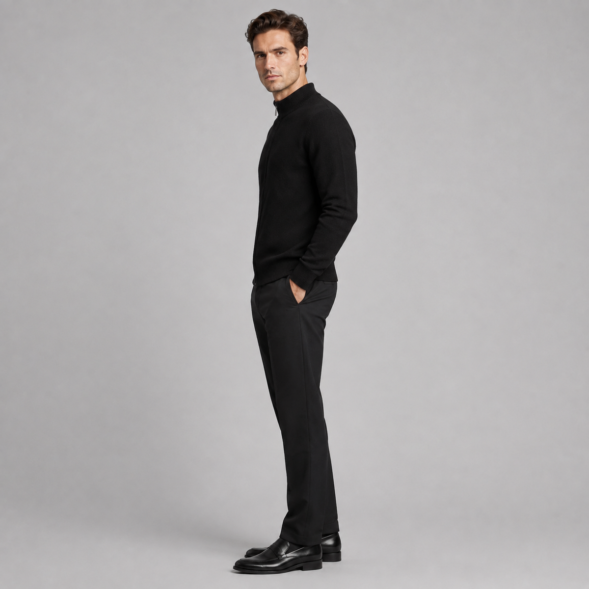 A man in a black mock-neck knit sweater and black trousers stands in profile against a light gray studio background, wearing black leather dress shoes.