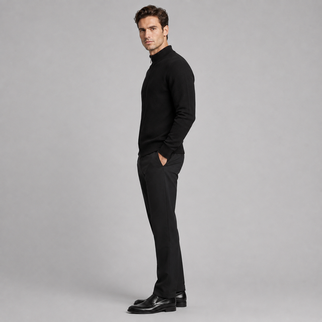 A man in a black mock-neck knit sweater and black trousers stands in profile against a light gray studio background, wearing black leather dress shoes.