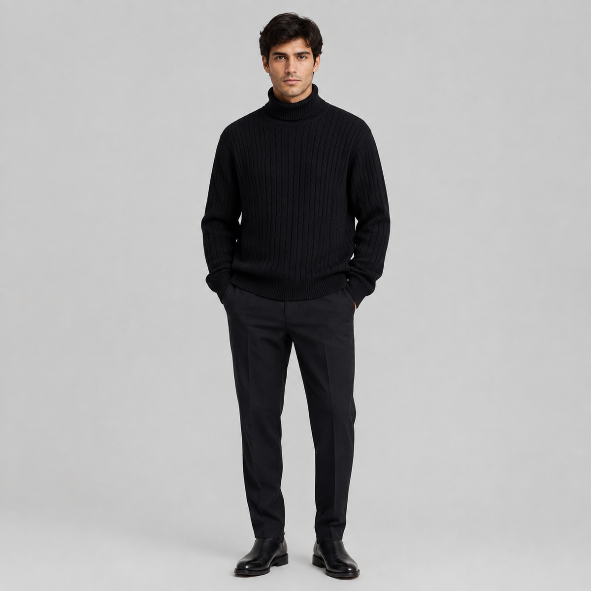 A man wearing a black ribbed turtleneck sweater, black trousers, and black dress shoes, standing against a light gray studio background.