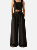 Person wearing a black sleeveless top and wide-leg pants with button details on a beige background