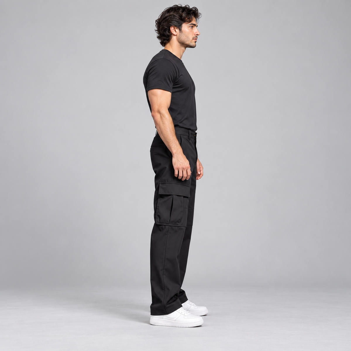 A man in a black t-shirt and black cargo pants stands in profile against a light gray studio background, wearing white sneakers.