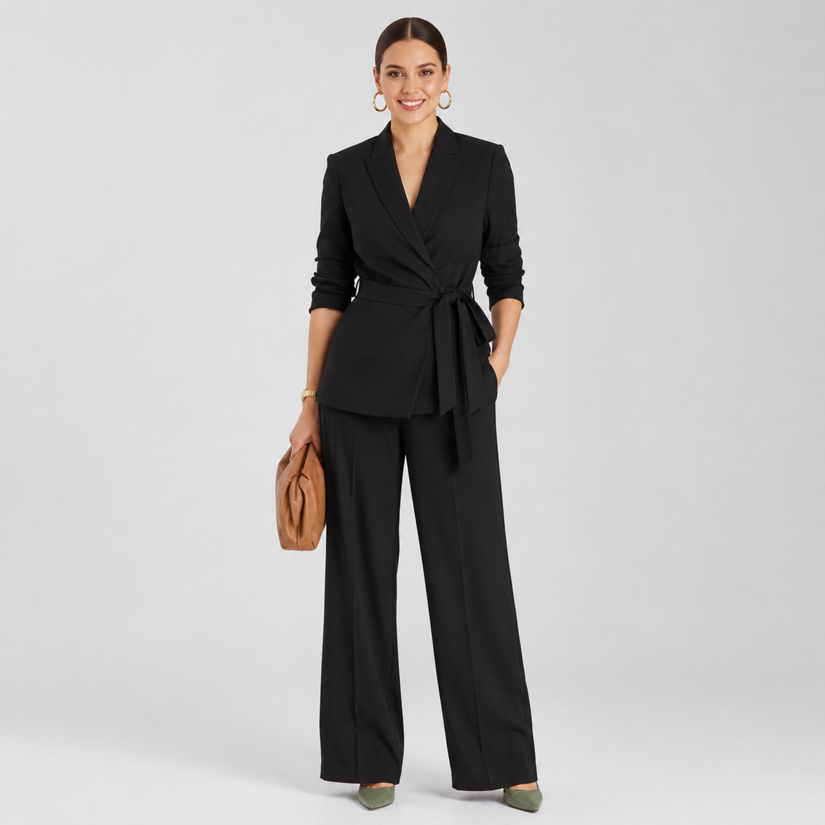 A woman modeling a black tailored two-piece outfit consisting of a wrap-style blazer with a tie waist and matching wide-leg trousers, photographed against a light gray studio background. She is holding a tan clutch and wearing olive green pointed shoes.
