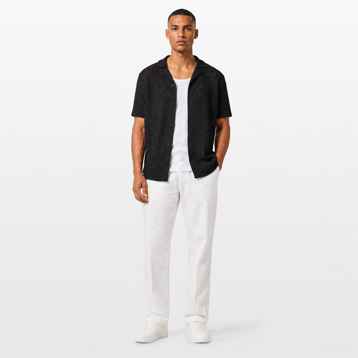 Man wearing a black textured shirt over a white shirt, paired with white pants on a white background