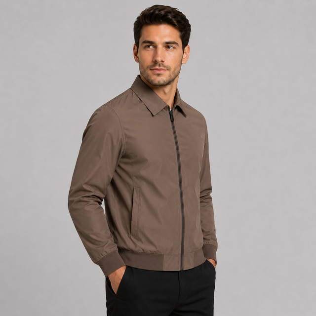 A man wearing a brown zip-up jacket with a collar and front pockets, paired with black pants, standing against a solid gray studio background.