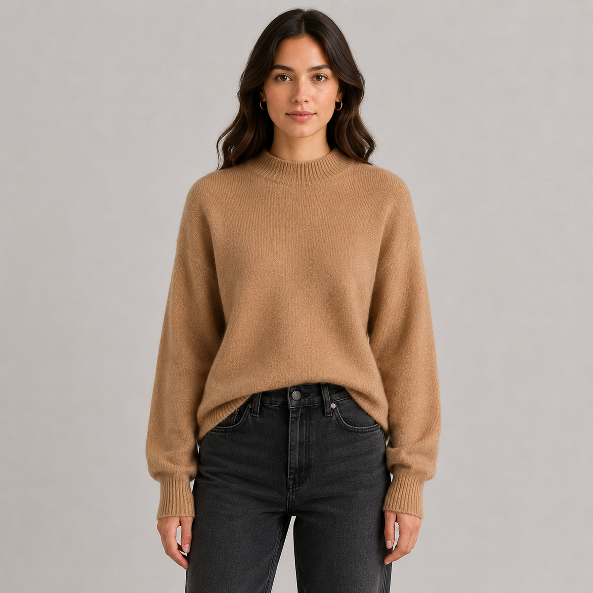 A woman wearing a camel-colored knit sweater and dark gray jeans, standing against a solid light gray studio background.