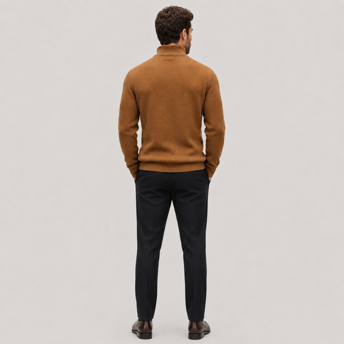 A man viewed from behind wearing a camel brown ribbed knit mock neck sweater, dark navy trousers, and brown leather dress shoes, standing against a light gray studio background.