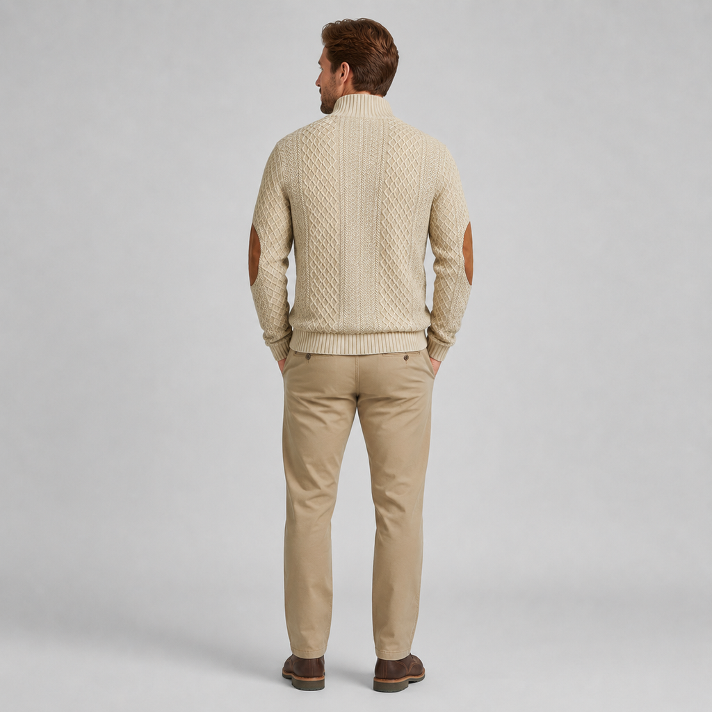 A man viewed from behind wearing a cream cable-knit sweater with brown leather elbow patches, paired with khaki pants and brown shoes, against a light gray studio background.
