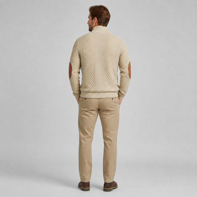A man viewed from behind wearing a cream cable-knit sweater with brown leather elbow patches, paired with khaki pants and brown shoes, against a light gray studio background.