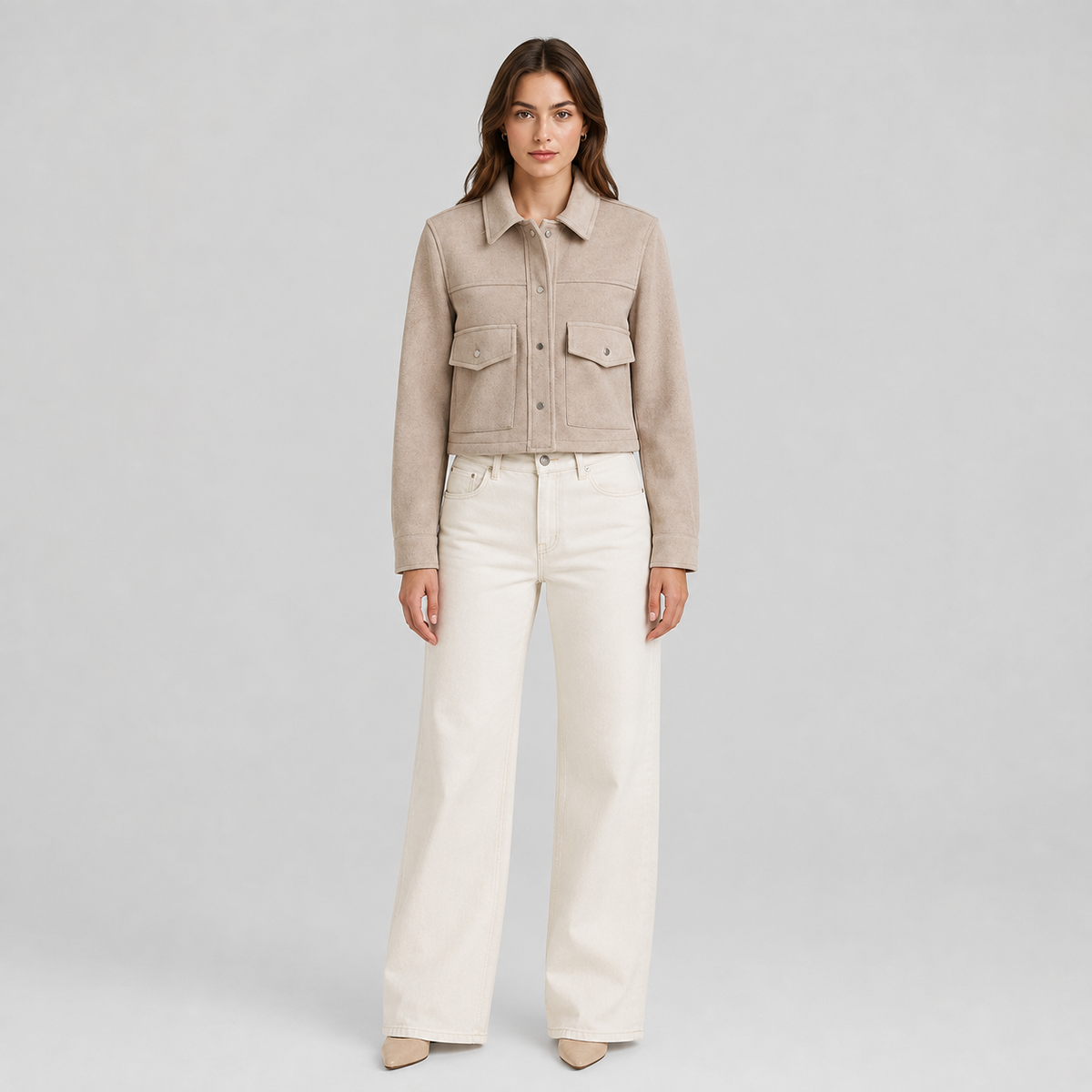 A woman stands against a light gray studio background wearing a cropped beige suede-like jacket with flap chest pockets and off-white wide-leg pants.