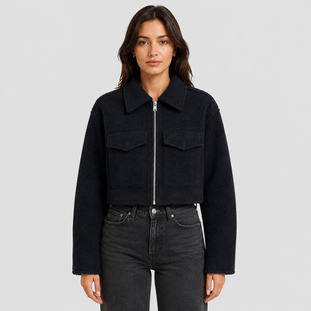 A woman modeling a cropped black fleece jacket with a silver front zipper and two chest flap pockets, paired with dark gray jeans, against a light gray studio background.