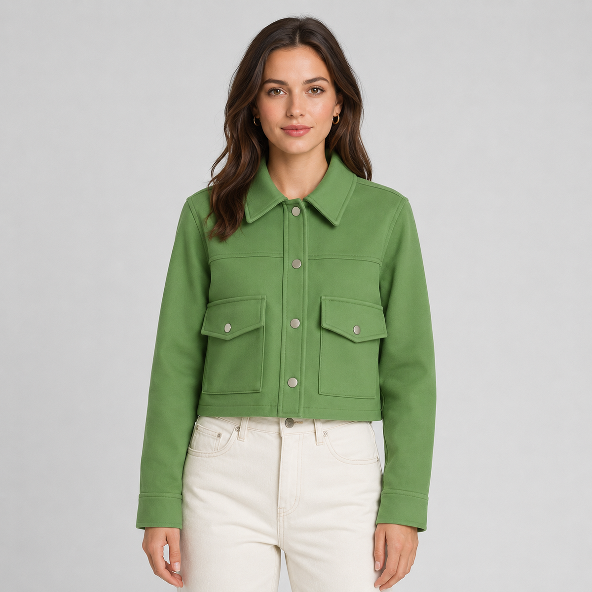 A woman modeling a cropped green button-front jacket with two flap chest pockets, paired with off-white jeans, against a light gray studio background.