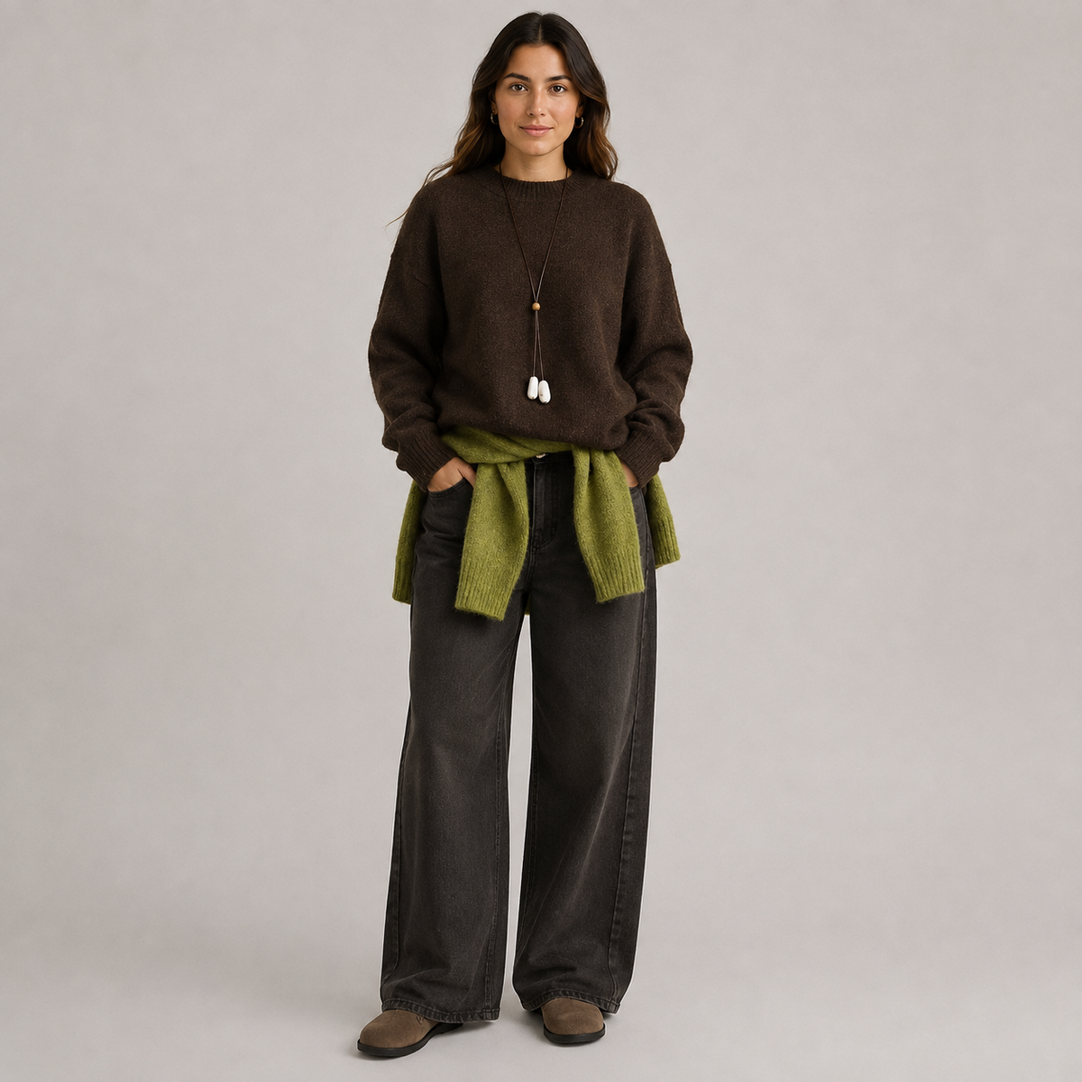 A woman stands against a plain light gray studio background wearing a dark brown knit sweater, wide-leg charcoal gray pants, and brown shoes. She has an olive green knit wrap tied around her waist and a long pendant necklace with white tassel charms.
