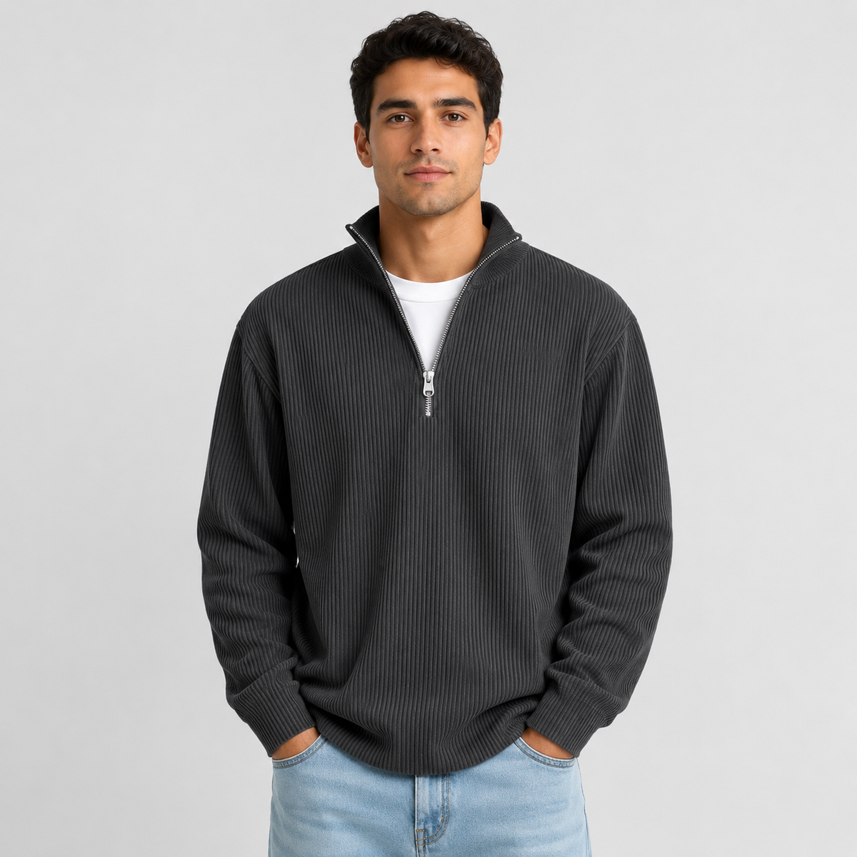 A man wearing a dark gray ribbed quarter-zip pullover sweater over a white t-shirt and light blue jeans, standing against a light gray studio background.