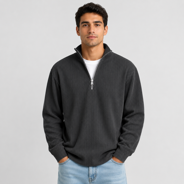 A man wearing a dark gray ribbed quarter-zip pullover sweater over a white t-shirt and light blue jeans, standing against a light gray studio background.