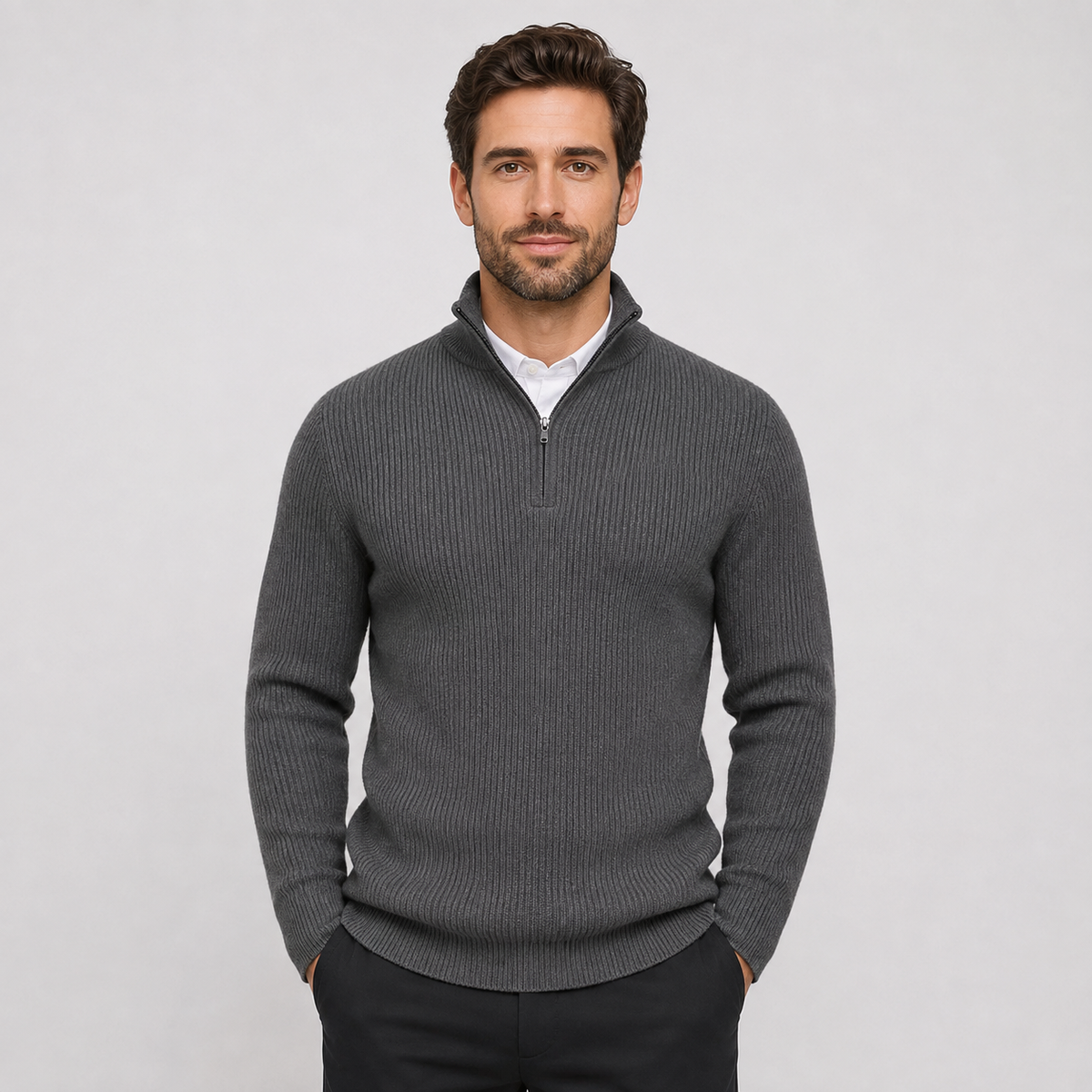 A man wearing a dark gray ribbed quarter-zip sweater over a white collared shirt and dark trousers, standing against a light gray studio background.