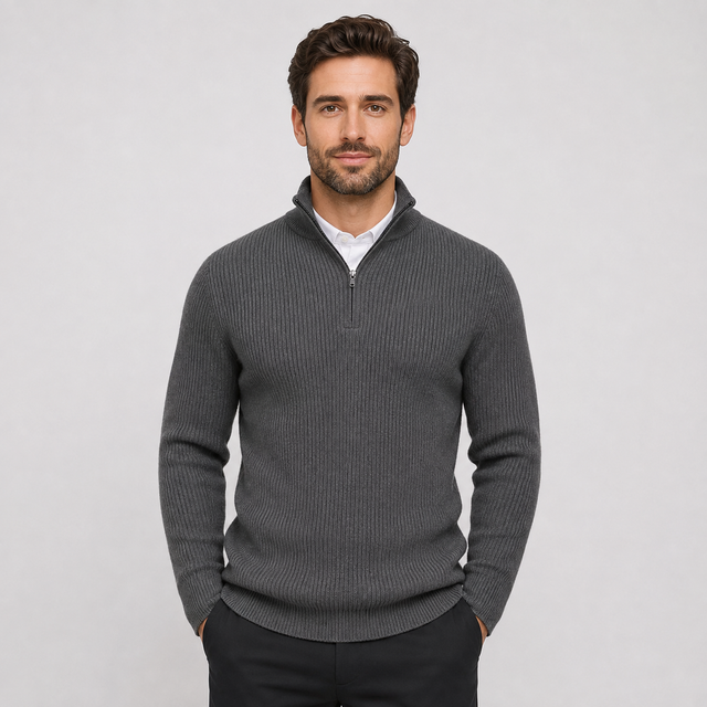 A man wearing a dark gray ribbed quarter-zip sweater over a white collared shirt and dark trousers, standing against a light gray studio background.