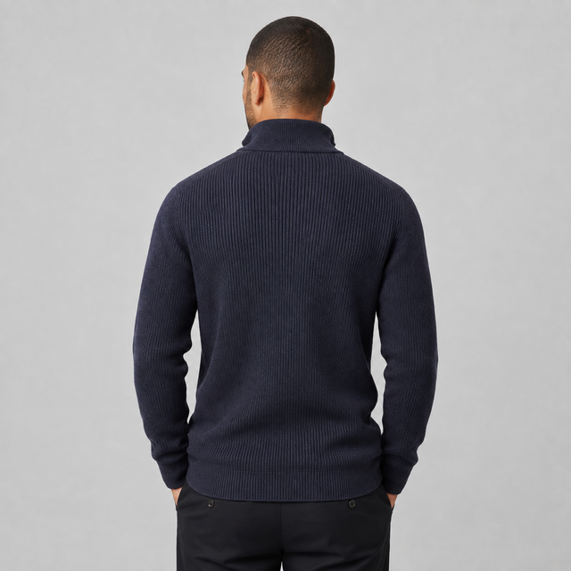 A man viewed from behind wearing a dark navy ribbed knit mock neck sweater and black trousers, standing against a neutral gray studio background.