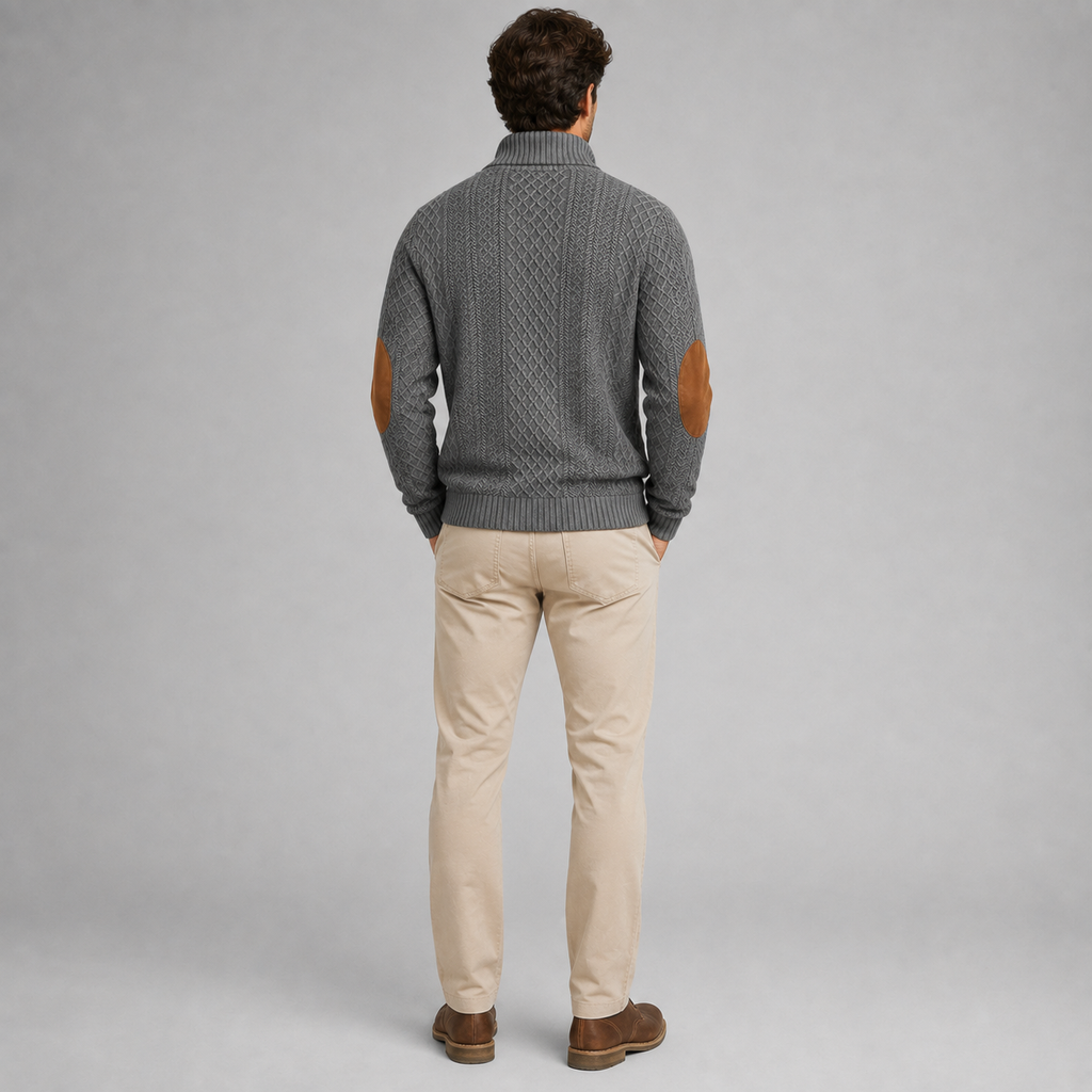 A man viewed from behind wearing a gray cable-knit sweater with brown leather elbow patches, paired with beige trousers and brown shoes, against a solid gray studio background.