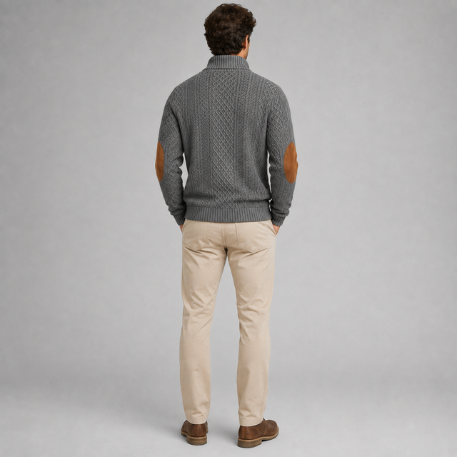 A man viewed from behind wearing a gray cable-knit sweater with brown leather elbow patches, paired with beige trousers and brown shoes, against a solid gray studio background.