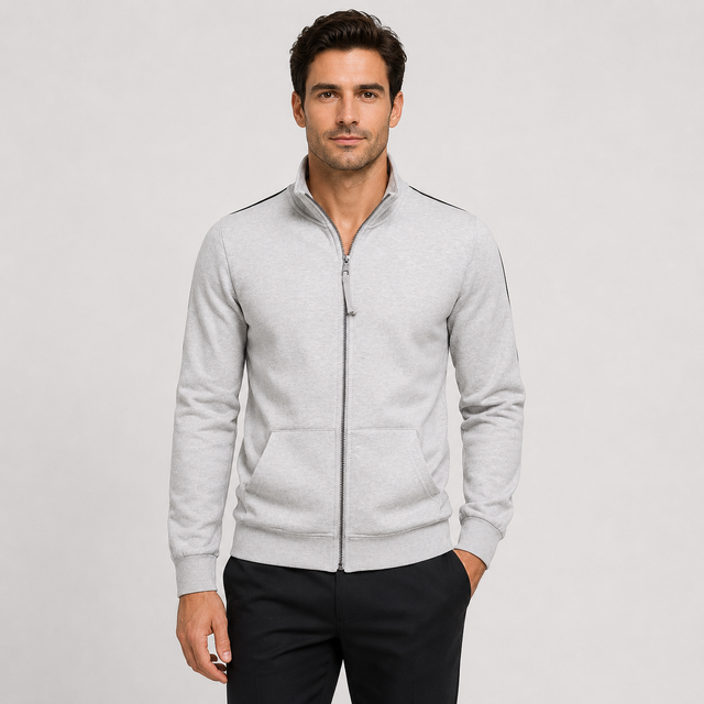A man wearing a light gray heather zip-up track jacket with black shoulder stripes and black pants, standing against a light gray studio background.