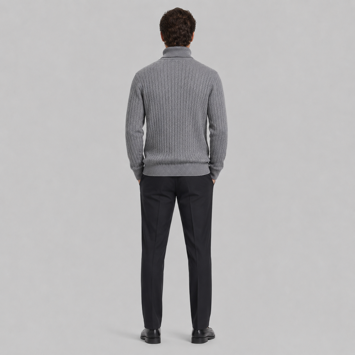 A man viewed from behind wearing a gray ribbed knit mock-neck sweater and dark trousers, standing against a light gray studio background.
