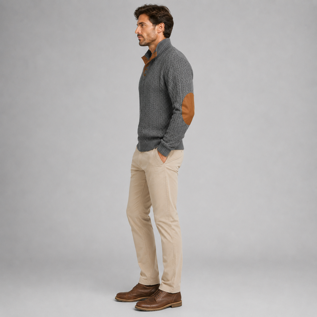 A man wearing a gray ribbed knit mock-neck sweater with brown elbow patches, paired with beige corduroy trousers and brown leather shoes, standing in profile against a light gray studio background.