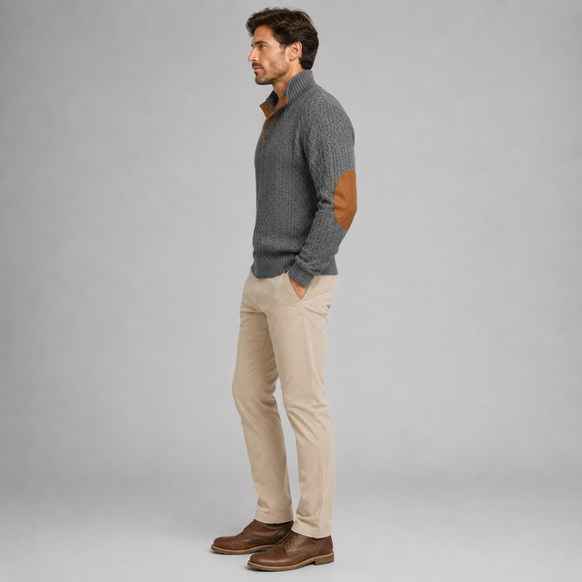 A man wearing a gray ribbed knit mock-neck sweater with brown elbow patches, paired with beige corduroy trousers and brown leather shoes, standing in profile against a light gray studio background.