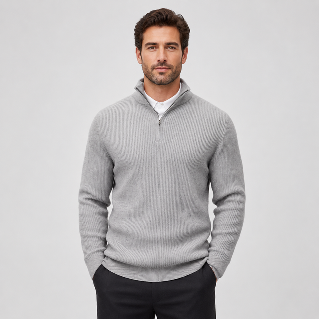 A man wearing a light gray ribbed quarter-zip sweater over a white collared shirt and dark trousers, standing against a plain light gray studio background.
