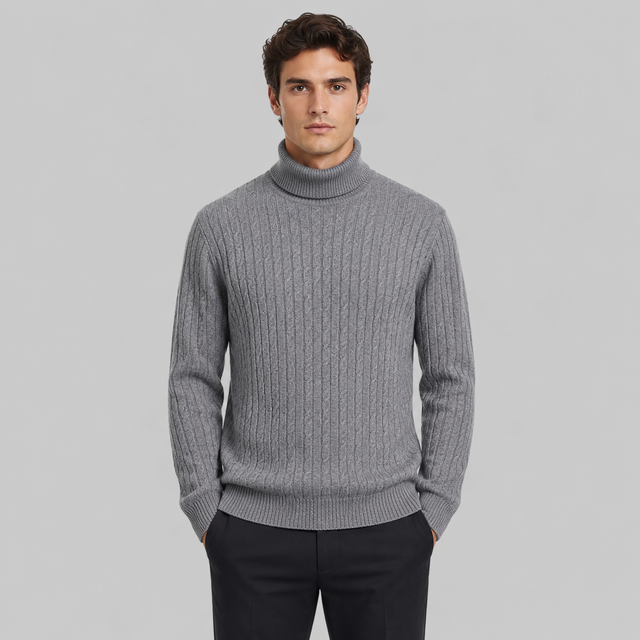 A man wearing a gray ribbed turtleneck sweater and dark trousers, standing against a solid light gray background.