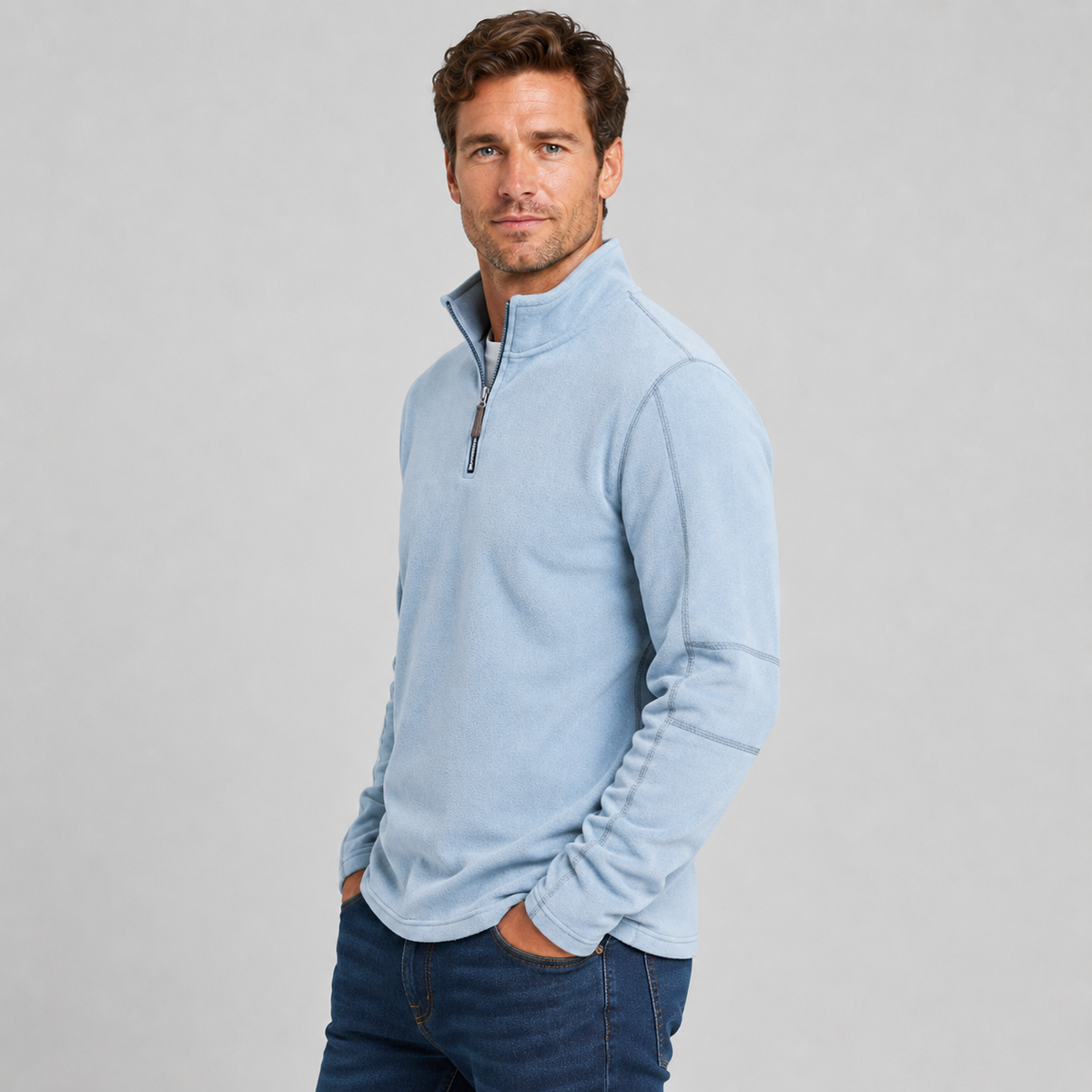 A man wearing a light blue quarter-zip fleece pullover sweater and dark blue jeans, standing against a neutral light gray studio background.