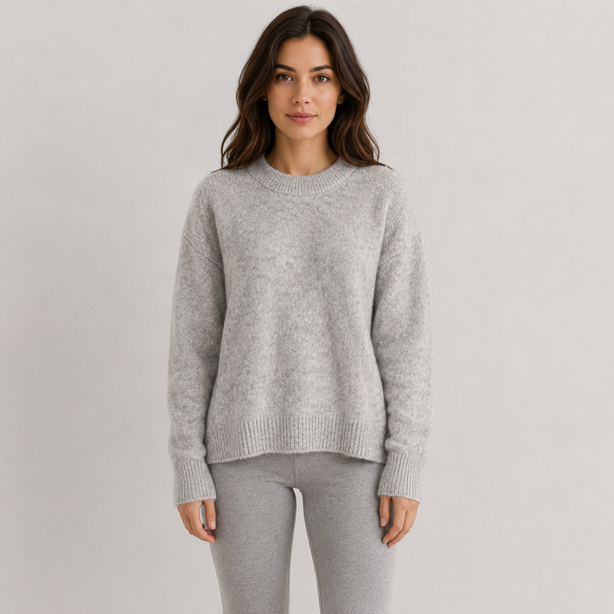 A woman wearing a light gray heathered knit crewneck sweater and matching gray pants, standing against a plain light gray studio background.