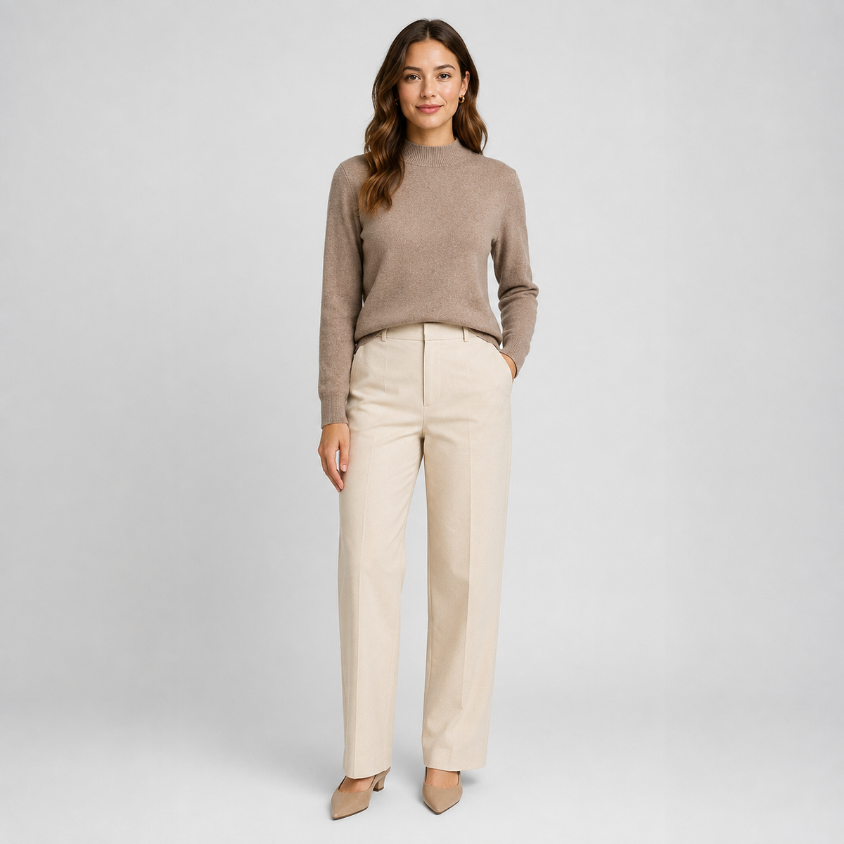 A woman stands against a light gray studio background wearing a taupe mock-neck knit sweater, straight-leg cream trousers, and beige pointed-toe heels.