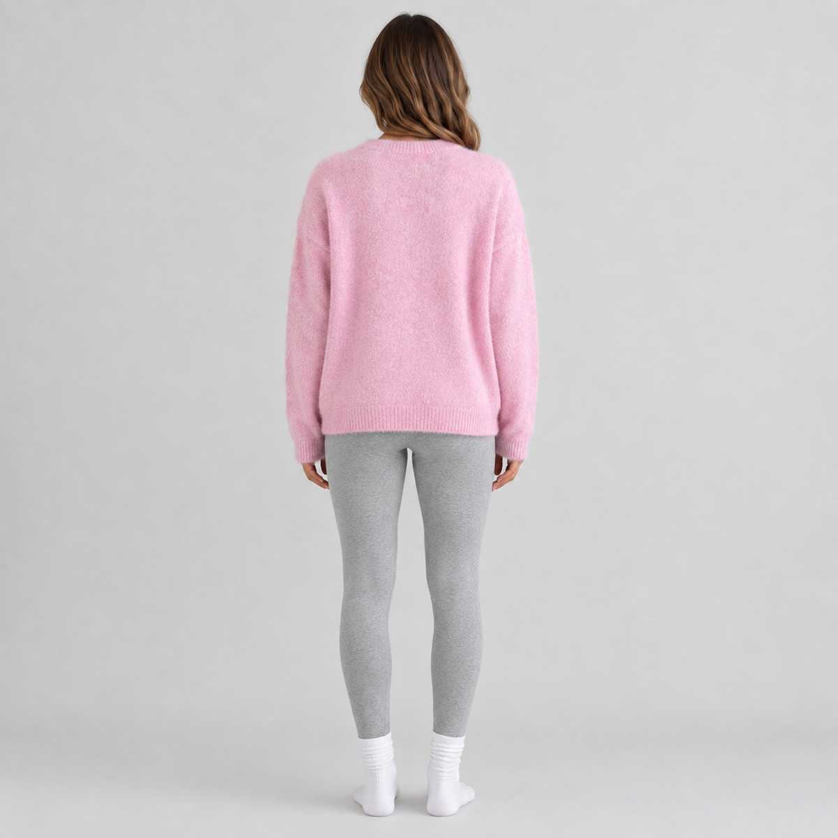 A woman viewed from behind wearing a fuzzy light pink crewneck sweater, light gray leggings, and white socks, standing against a neutral light gray studio background.