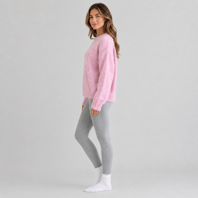 A woman modeling a fuzzy light pink long-sleeve sweater paired with heather gray leggings and white socks, standing against a light gray studio background.