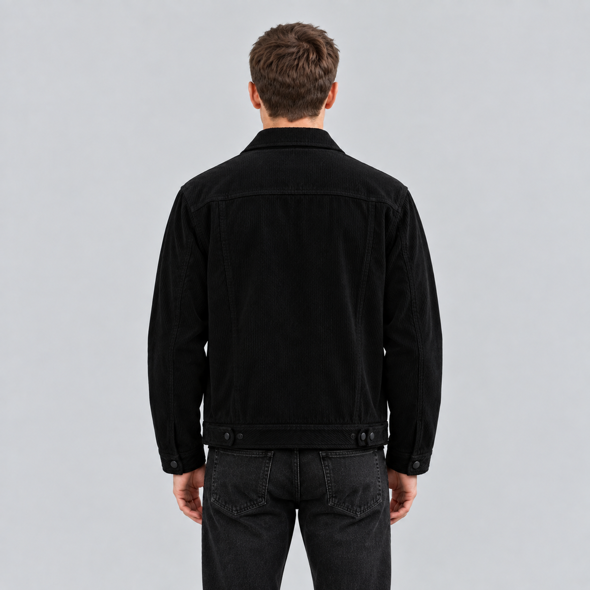 A man viewed from behind wearing a black corduroy jacket and dark gray jeans against a light gray studio background.