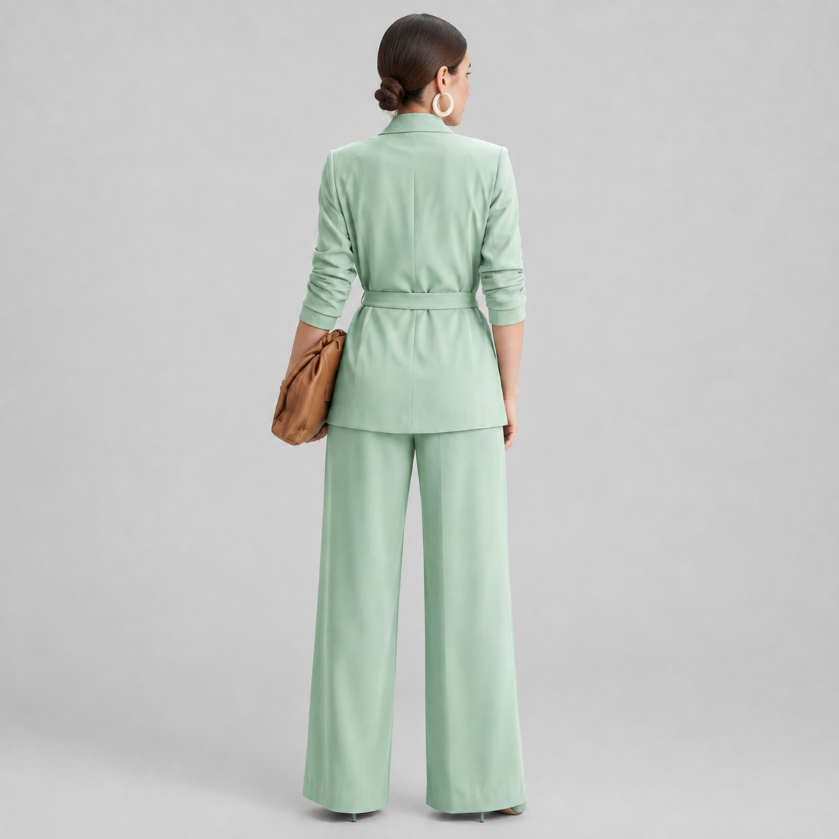 A woman shown from the back wearing a light mint green tailored set consisting of a long-sleeve belted blazer and matching wide-leg trousers, against a plain light gray studio background.