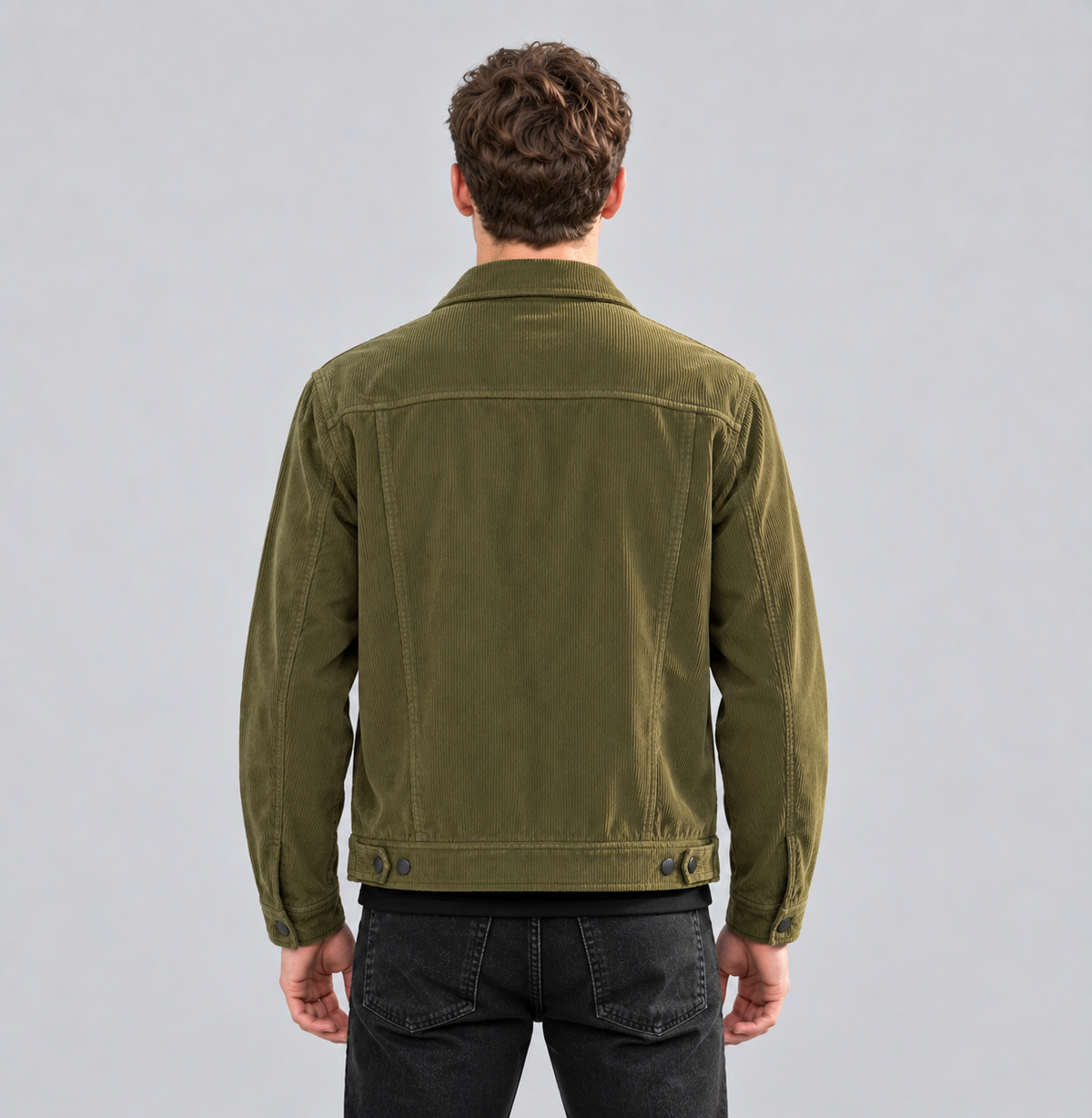 A man viewed from behind wearing an olive green corduroy jacket and black jeans, standing against a solid gray studio background.