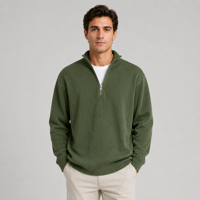 A man wearing an olive green ribbed quarter-zip sweater over a white t-shirt and beige pants, standing against a solid light gray background.